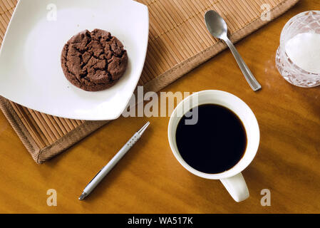 Coffee break - american coffee in ceramic white cup, cookie on white plate, teaspoon, pen, placemat and sugar-bowl on wooden table, messy arrangement Stock Photo