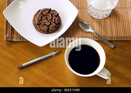 Black coffee in ceramic white cup, chocolate cookie on white plate, teaspoon, pen, bamboo placemat and sugar-bowl on wooden table, indoors, top view Stock Photo