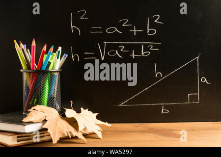 Books, a pencil holder with pencils, pens and various school supplies, and some autumn leaves on wooden boards with a blackboard in the background wit Stock Photo