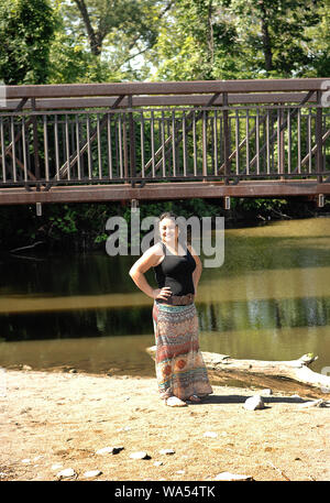 A happy young woman in a long dress standing in the park under an old bridge on a small lake on a nice sunny day Stock Photo