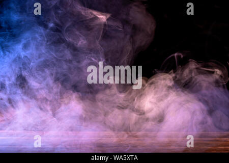Conceptual image of multi-colored smoke isolated on dark black background and wooden table. Stock Photo