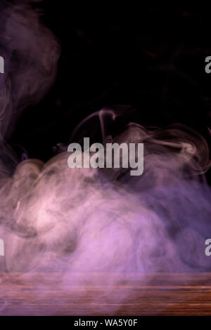Conceptual image of multi-colored smoke isolated on dark black background and wooden table. Stock Photo