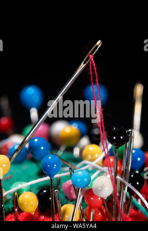 Close up of sewing tools.  Needle and red thread stuck in pin cushion with many colorful stick pins, all on black background; Stock Photo