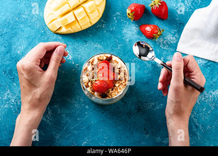 top view hands of girl with spoon eating dessert with mango, granola, curd mousse and strawberries on blue background. concept of healthy desserts Stock Photo
