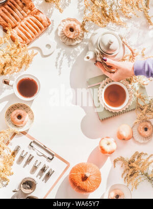 Top view of cozy autumn concept. Female hand holding teapot on white background with mini donuts and pastry, tea cups, flowers,clipboard and pumpkin. Stock Photo