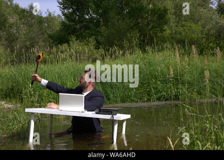 young hipster bearded guy lounges, distracted from work in the office and taking a selfie. At a table standing in a swamp in the water Stock Photo