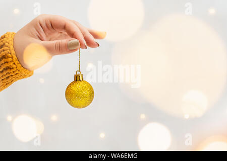 Woman hand in orange sweater holding golden Christmas tree ball with garland bokeh on background. Stock Photo