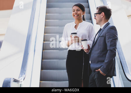 Businessman and businesswoman standing together on escalator and talk about business holding coffee in hand Stock Photo