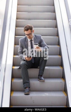 Businessman sitting on escalator surfing the net and holding coffee in hand Stock Photo