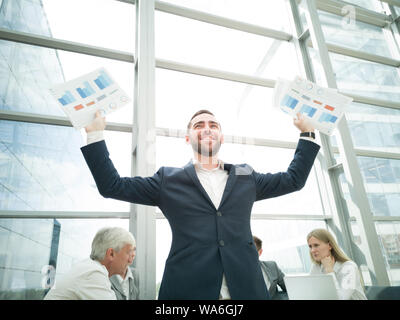 Young joyful businessman standing with arms raised holding data statistics diagrams and smiling , team of his colleagues sitting at the table in the b Stock Photo