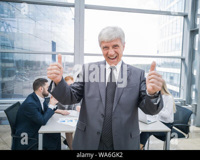 Confident mature businessman showing thumb up and smiling , team of his colleagues sitting at the table in the background Stock Photo
