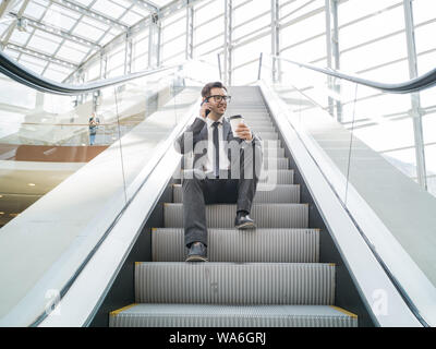 Businessman sitting on escalator talking on phone and holding coffee in hand Stock Photo