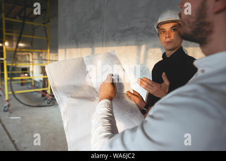 Safety inspector and build worker inside building under construction. Engineer hold blueprint in hand and discussing project with builder on construct Stock Photo