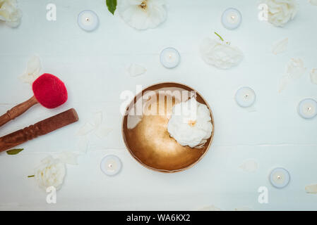 Top view Tibetan singing bowl with floating in water rose inside. Special sticks, burning candles, flowers petals on the white wooden background. Medi Stock Photo