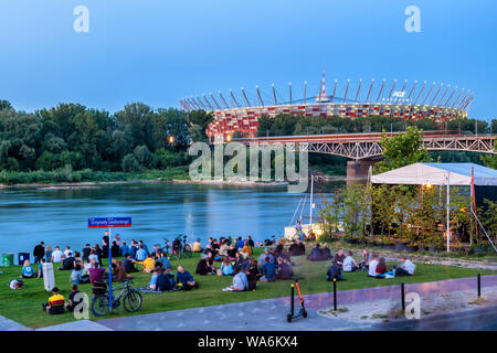 Warsaw, Poland - July 18, 2019: People relax by the Vistula River with view to the National Stadium illuminated at dusk Stock Photo