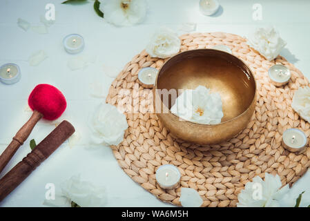 Tibetan singing bowl with floating in water rose inside. Special sticks, burning candles, flowers petals on the white wooden background. Meditation an Stock Photo