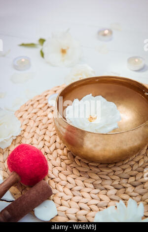 Tibetan singing bowl with floating in water rose inside. Special sticks, burning candles, flowers petals on the white wooden background. Meditation an Stock Photo