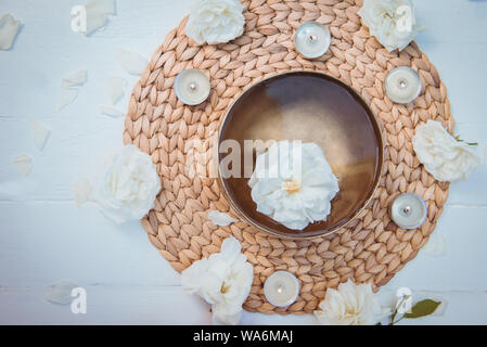 Close up Top view Tibetan singing bowl with floating in water rose inside. Burning candles, flowers petals on the white wooden background. Meditation Stock Photo