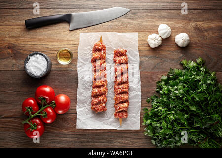 Raw Turkish Adana shish kebab on white cooking paper and wooden table. Decorated with herbs, spices and knife. Overhead view. Stock Photo