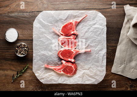 Slices of raw lamb rib chops on white cooking paper and wooden table. Decorated with salt, pepper, meat fork and napkin. Overhead view. Stock Photo