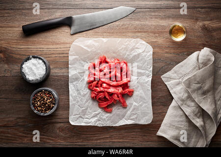 Chopped raw veal meat for beef stroganoff on white cooking paper and wooden table. Decorated with salt, pepper, meat fork and napkin. Overhead view. Stock Photo