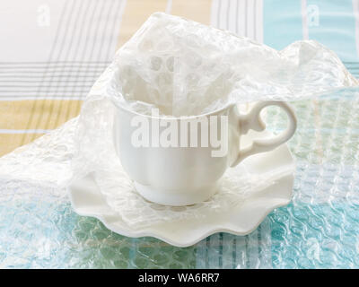 White elegant porcelain tea cup and saucer half packaged with transparent bubble wrap on a checkered tablecloth. Material for packing fragile items. Stock Photo