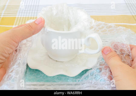 Woman hands package white elegant tea cup and saucer with transparent bubble wrap on a checkered tablecloth. Material for packing fragile items. Stock Photo