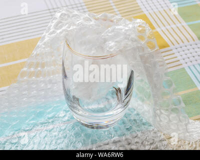 Thick bottom glass half packaged with transparent bubble wrap on a checkered tablecloth. Material for packing fragile items for safe transportation. Stock Photo