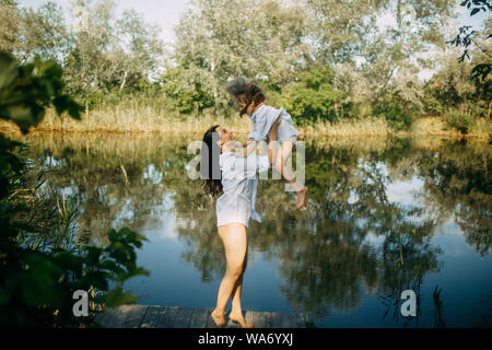 Mother and daughter play and have fun on a wooden bridge near a small river with reflection of trees and sky in the water. Stock Photo