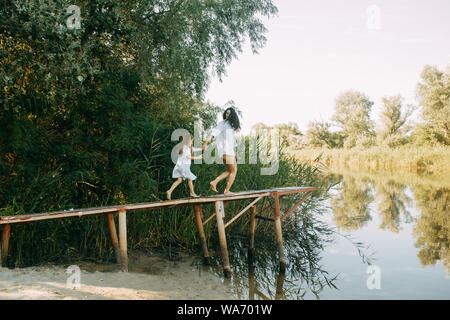Mother and daughter are running and having fun on a wooden bridge on background of river and reeds. Stock Photo