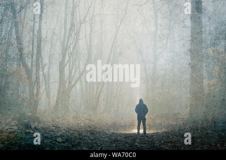 An artistic, double exposure of a hooded man standing in a spooky forest on a foggy winters day. With a grunge, blurred edit. Stock Photo