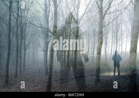 An artistic double exposure of a hooded figure standing in a forest on a spooky, foggy winters day. Looking at a man with a torch. Stock Photo