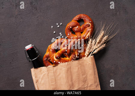 Fresh salted pretzels and beer bottle in the paper bag on the brown table decorated with wheat ears, top view composition Stock Photo
