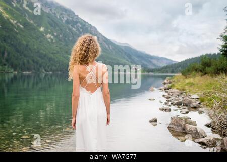 Beautiful young woman with curly hair in a white dress stands near incredible lake. Tatra National park, Poland. Famous lake Morskie Oko or Sea Eye Stock Photo