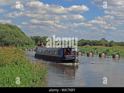 A canal narrow boat is navigating along the canalised river Trent near Alrewas, passing a weir barrier and pushing against the flow of the river. Stock Photo