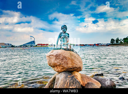 Statue of little mermaid sitting on stone. Behind it is the sea and the view of Copenhagen harbour.
