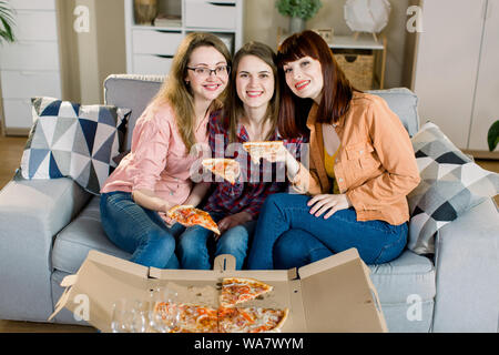 Portrait of three happy young female friends in casual clothes eating pizza on sofa at home. Women friendship, eating together Stock Photo