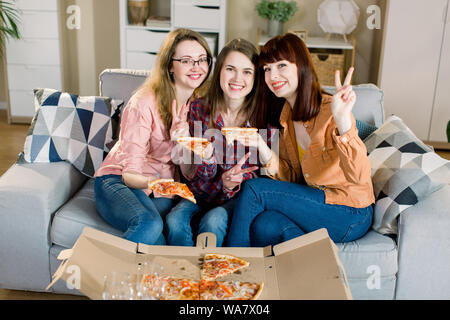 Portrait of three happy young female friends in casual clothes eating pizza on sofa at home. Women friendship, eating together Stock Photo
