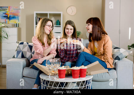 friendship, people, pajama party and junk food concept - happy young three women or girls eating pizza, popcorn and drinking wine at home Stock Photo