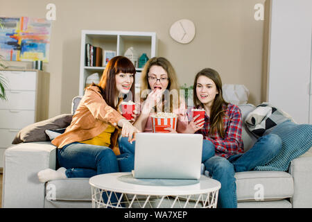 Food and movies. Three emotional pretty sisters eating popcorn when watching movie or sport program while sits on the sofa of beautiful room at home