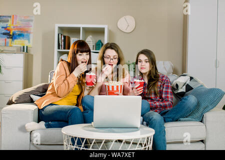 Food and movies. Three emotional pretty sisters eating popcorn when watching movie or sport program while sits on the sofa of beautiful room at home