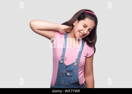 Neck pain. Portrait of sad brunette young girl in pink t-shirt and blue overalls standing and holding her painful neck and feeling bad. indoor studio Stock Photo