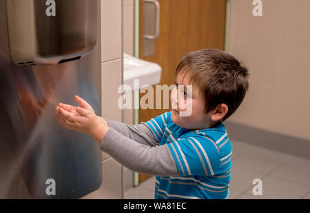 A young boy uses a air hand dryer to dry his hands after washing them in the bathroom sink. Stock Photo