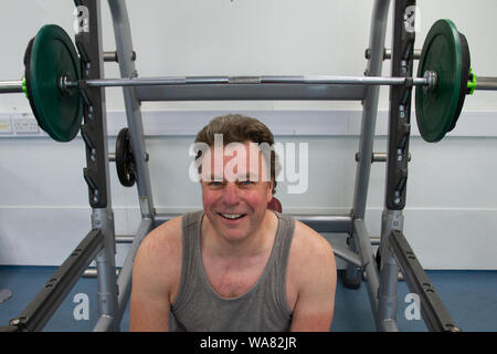 An head and shoulders portrait of a man smiling for the camera as he sits in front of  weights in a gym in between exercise. Stock Photo