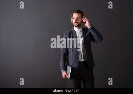 A serious handsome young businessman in a suit holding a tablet and listening to music in front of a dark grey background in a studio. Stock Photo