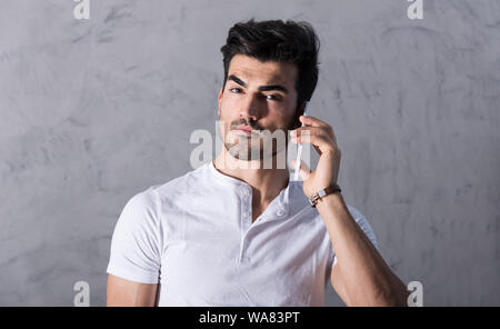 A serious handsome young man in a white tshirt talking on his phone in front of a grey wall in a studio. Stock Photo