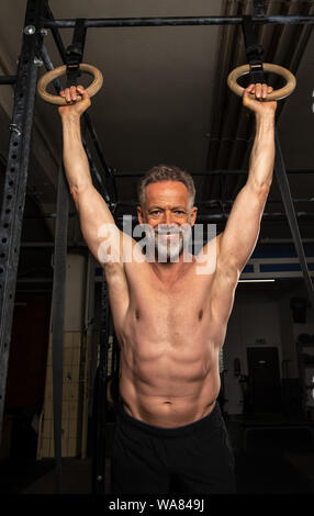 Portrait of an attractive older male athlete with strong muscles. The muscular and smiling bearded man is doing an exercise with the rings. Stock Photo