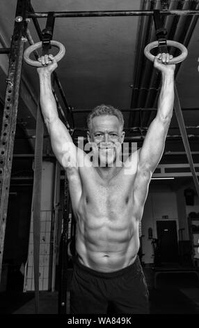 Portrait of an attractive older male athlete with strong muscles. The athletic and smiling bearded man is doing an exercise with the rings. Stock Photo