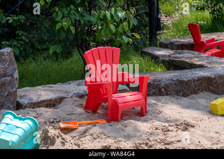 small child's Adirondack chair rests in a big outdoor sand box with plenty of sand toys Stock Photo