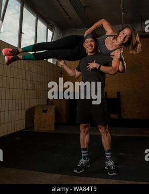 A muscular beautiful couple is smiling, doing a fitness workout together and having fun.The strong man is holding the fit woman on his shoulders. Stock Photo